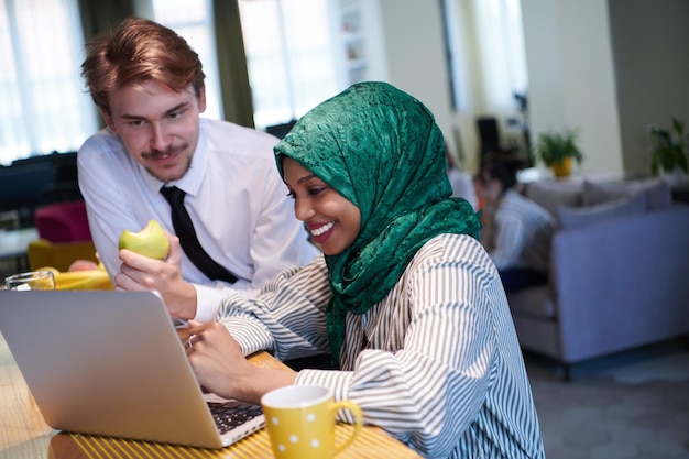 equipe de negócios multiculturais internacionais homem comendo maçã mulher muçulmana africana vestindo hijab verde bebendo chá enquanto trabalhava juntos no computador portátil na área de relaxamento no plano aberto moderno startu