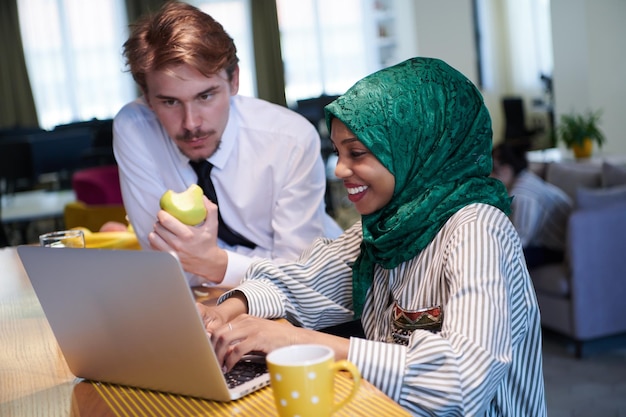 equipe de negócios multiculturais internacionais homem comendo maçã mulher muçulmana africana vestindo hijab verde bebendo chá enquanto trabalhava juntos no computador portátil na área de relaxamento no plano aberto moderno startu