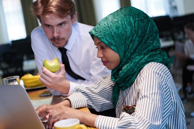 equipe de negócios multiculturais internacionais homem comendo maçã mulher muçulmana africana vestindo hijab verde bebendo chá enquanto trabalhava juntos no computador portátil na área de relaxamento no plano aberto moderno startu