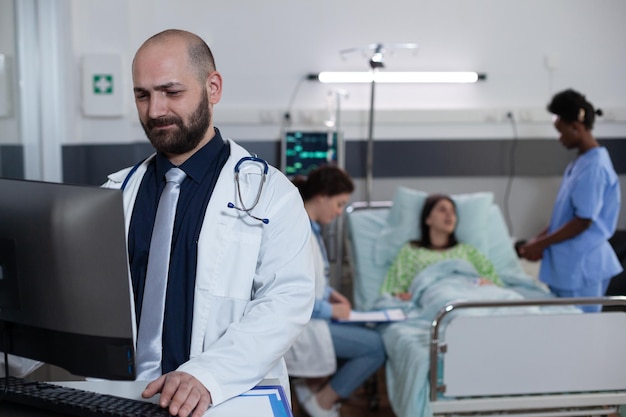 Equipe de profissionais de saúde que consulta o paciente com doença na cama do hospital enquanto o médico com estetoscópio está lendo os resultados do laboratório na tela do computador. Médico investigando registros médicos.