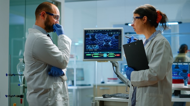 Equipe de químicos trabalhando com imagem de varredura de DNA olhando para a mesa de pé no laboratório de pesquisa médica, analisando amostras de bioquímicos, conversando. Desenvolvimento de microbiologia com equipamentos avançados