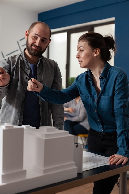 Foto equipe de trabalho do arquiteto falando no escritório profissional em pé na mesa com o modelo de construção da maquete. parceiros de projeto caucasianos em busca de um projeto de layout de construção