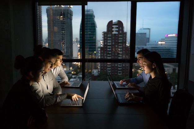 Equipe do negócio que trabalha tarde da noite com as luzes apagadas e computadores laptop tela acesa.