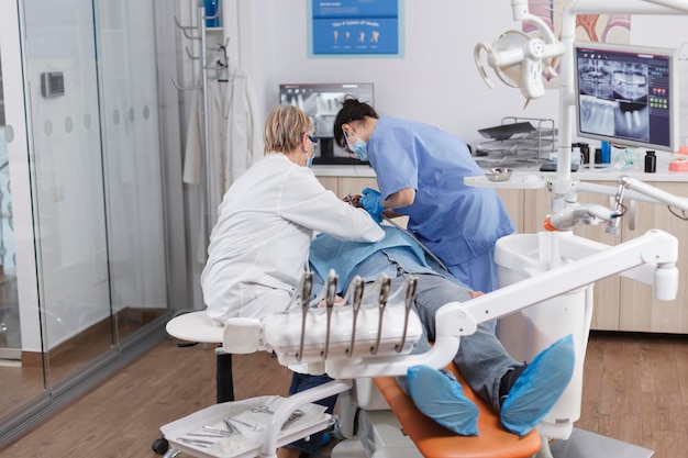 Equipe especializada de estomatologistas com máscara facial médica durante a cirurgia estomatológica na sala do consultório odontológico. Enfermeira ortodontista usando instrumento de broca discutindo tratamento de infecção com médico sênior