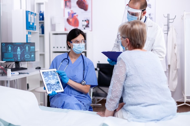 Equipe médica explicando o diagnóstico a uma idosa em um quarto de hospital usando um tablet e uma máscara facial contra o coronavírus como medida de segurança