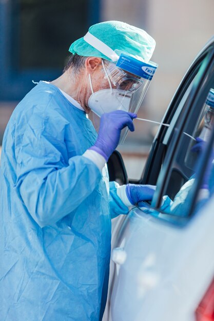 Equipe médica usando um EPI, realizando PCR com um cotonete na mão, em um paciente dentro de seu carro para detectar se ele está infectado com COVID-19