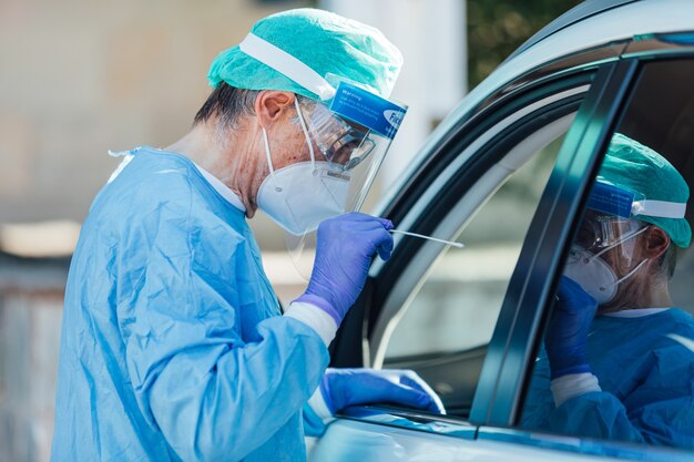 Equipe médica usando um EPI, realizando PCR com um cotonete na mão, em um paciente dentro de seu carro para detectar se ele está infectado com COVID-19