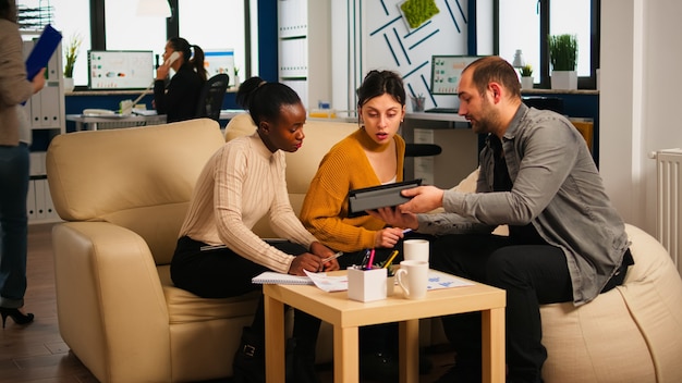 Equipe multiétnica analisando informações do tablet discutindo sentado no sofá brainstorming sobre um novo projeto para uma empresa start up. Diversos empresários analisando relatórios financeiros durante a reunião.
