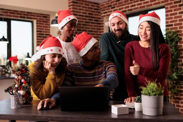 Equipe multiétnica de pessoas usando laptop no escritório festivo, trabalhando em negócios de inicialização durante a temporada de férias de natal. Fazendo trabalho em equipe no local de trabalho decorado com enfeites de natal.