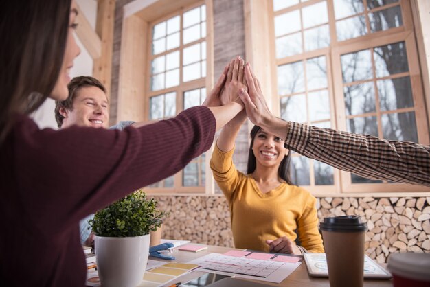 Equipe perfeita. Close de high five de pessoas positivamente agradáveis e encantadas, sentados juntos ao redor da mesa