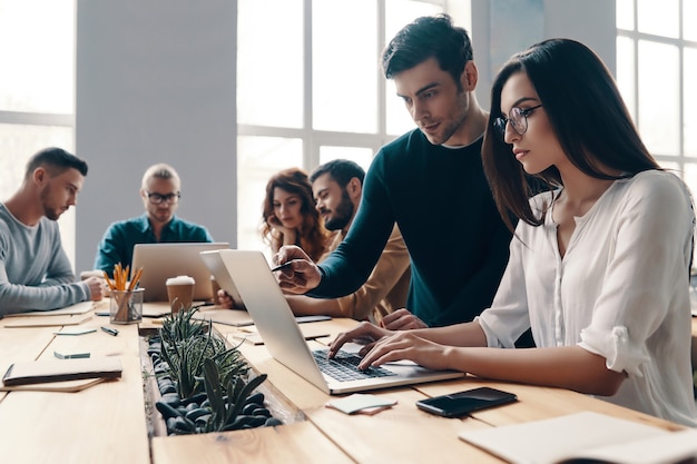 Equipe trabalhando junto. Grupo de jovens modernos em roupas casuais inteligentes, usando tecnologias modernas, enquanto trabalham no escritório criativo