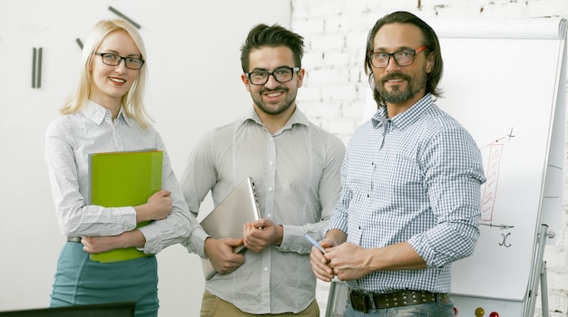 Foto equipo acertado del negocio toothy que sonríe mientras que se opone a la pared y al whiteboard blancos de la oficina en la presentación.