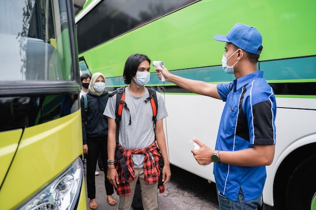 Un equipo de autobús con uniformes azules y un sombrero con una pistola térmica inspecciona al pasajero masculino con la máscara antes de abordar el autobús.