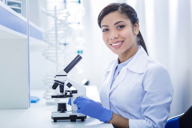 Equipo científico. Encantado de mujer inteligente inteligente sentada frente al microscopio y mirándote mientras haces una investigación biológica