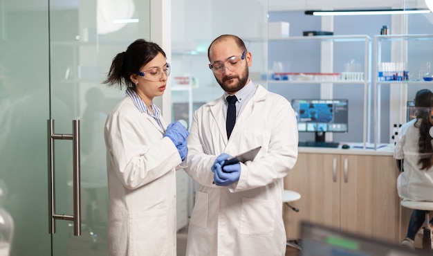 Equipo de científicos médicos que llevan a cabo el desarrollo de vacunas con la ayuda de tecnología de alta tecnología, médico sosteniendo una tableta analizando nueva información