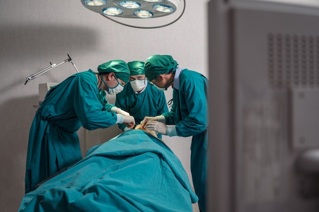 Equipo de cirujanos asiáticos que realizan una cirugía a un paciente gravemente herido a través de un monitor médico en el quirófano del hospital