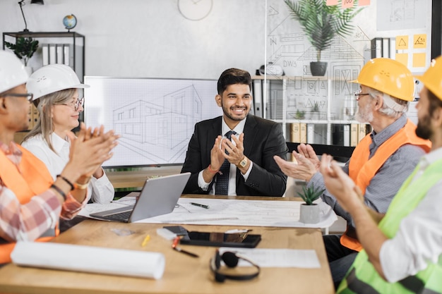 Equipo de construcción feliz sentado en la oficina y aplaudiendo durante la presentación del nuevo proyecto