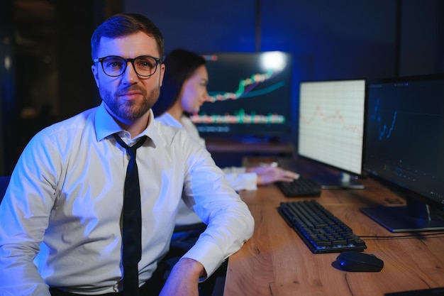 Un equipo de corredores de bolsa está teniendo una conversación en una oficina oscura con pantallas de visualización