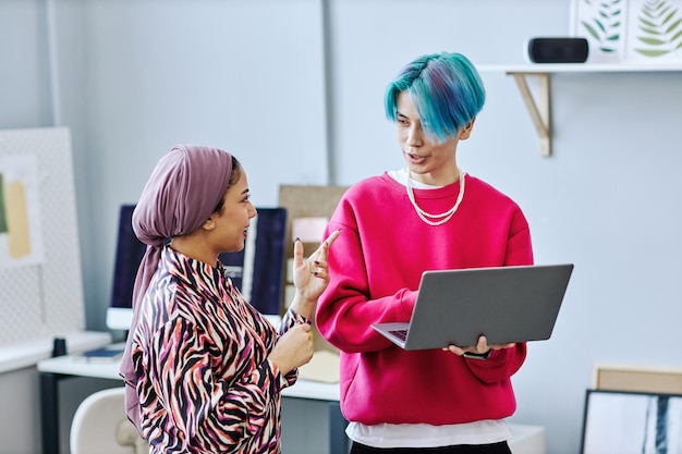 Equipo creativo de la generación Z discutiendo el proyecto de pie en la oficina y sosteniendo una computadora portátil