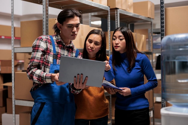 El equipo de empleados asiáticos del almacén postal analiza la lista de pedidos de los compradores en una computadora portátil. Trabajadores y trabajadoras del almacén que utilizan el software del sistema de seguimiento de paquetes y cooperan en la sala de almacenamiento