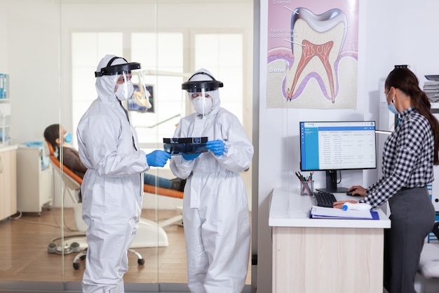 El equipo de estomatología se vistió con un traje de ppe durante la pandemia global con coronavirus en la recepción dental con una radiografía del paciente, manteniendo el distanciamiento social