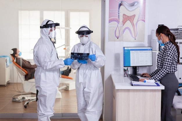 El equipo de estomatología se vistió con un traje de ppe durante la pandemia global con coronavirus en la recepción dental con una radiografía del paciente, manteniendo el distanciamiento social