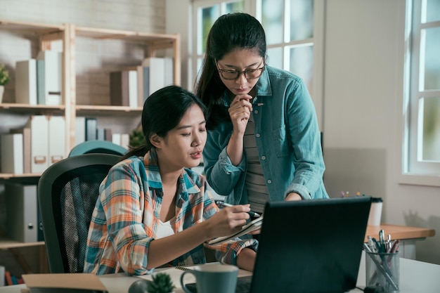 El equipo femenino asiático joven que trabaja en la puesta en marcha discute el proyecto frente a la computadora portátil. oficina de estudio moderna y luminosa. vintage dos mujeres japonesas en traje casual trabajando considerando una lluvia de ideas en interiores