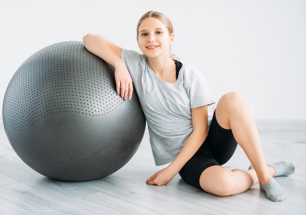 Equipo de fitness Deporte en el interior Niños activos Esenciales de entrenamiento Accesorios de gimnasia Sonriente niña atlética feliz sentada en el suelo con pelota de gimnasia sobre fondo claro