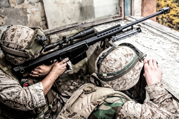 Equipo de francotiradores de los marines armado con un rifle de francotirador antimaterial de gran calibre escondido en un edificio urbano en ruinas, disparando objetivos enemigos a distancia desde el refugio, sentado en una emboscada. Tiroteo militar en la ciudad