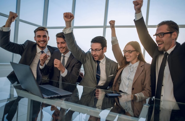 Foto equipo de hombres de negocios celebrando el éxito sentados en la computadora con las manos en los puños apretados y sonriendo mirando la pantalla del portátil en la mesa frente a ellos