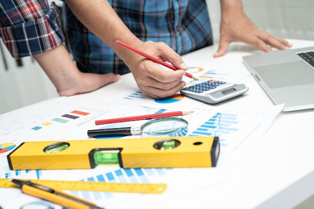 El equipo de ingenieros y socios asiáticos aprende y habla con el gráfico del proyecto del plan de la casa y el casco amarillo y herramientas de construcción de ingeniería en el trabajo de mesa