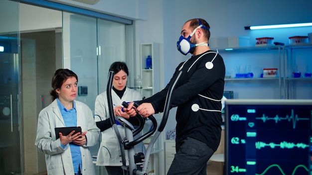 Equipo de investigadores médicos que monitorean el rendimiento del atleta con máscara y electrodos adheridos a él midiendo la resistencia, la frecuencia cardíaca, la resistencia psicológica, el entrenamiento de la masa muscular en el laboratorio de ciencias deportivas