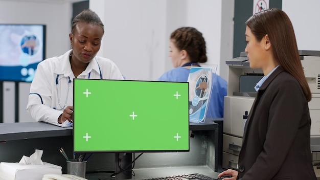 Equipo médico diverso que usa computadora con pantalla verde en el mostrador de recepción. El personal de la instalación trabaja con un fondo de espacio de copia aislado, una plantilla de maqueta en blanco y una pantalla de clave de croma en el monitor.