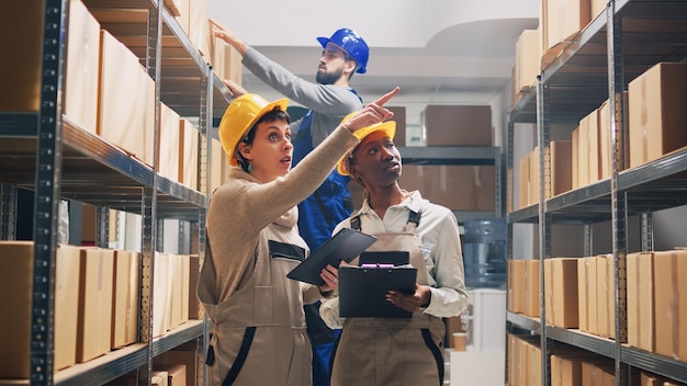 Equipo de mujeres diversas que planean el envío de mercancías, revisando productos en papeles. Supervisor de depósito mirando stock en tabletas y organizando mercancías, los paquetes de cartón funcionan.