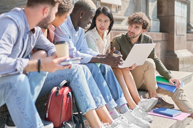 El equipo multicultural de estudiantes está charlando en los escalones del campus.