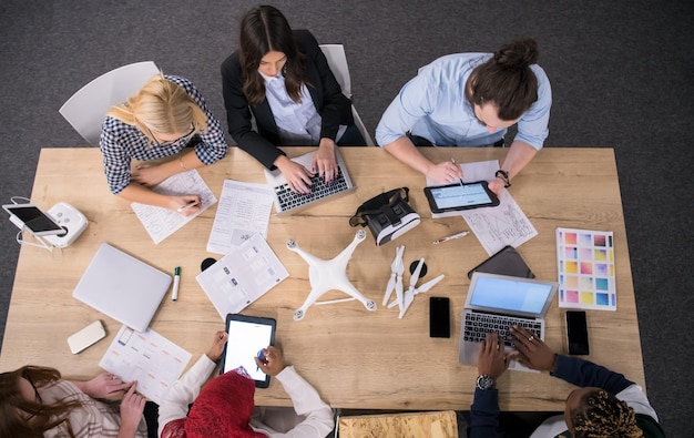 equipo multiétnico de empresas emergentes discutiendo un nuevo plan de negocios, trabajando en computadoras portátiles y tabletas mientras aprende sobre la tecnología de drones para futuras empresas comerciales vista superior