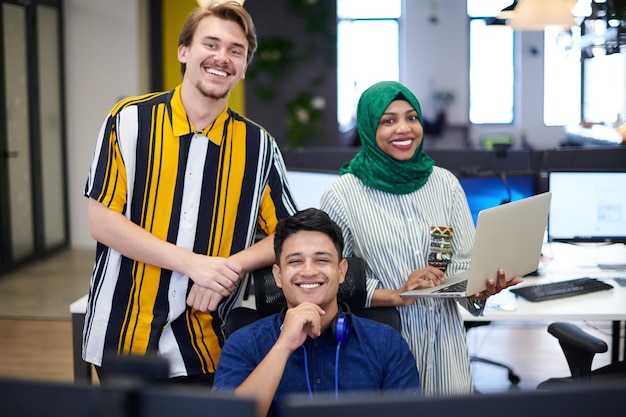 Equipo multiétnico de empresas emergentes Mujer árabe que usa un hiyab en una reunión en una moderna oficina de planta abierta con lluvia de ideas en el interior, trabajando en una computadora portátil y una computadora de escritorio