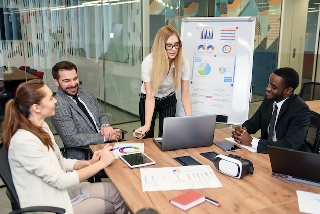 Equipo multiétnico profesional muy enérgico y confiado que trabaja junto con un proyecto empresarial conjunto en la sala de reuniones utilizando una computadora portátil e informes.