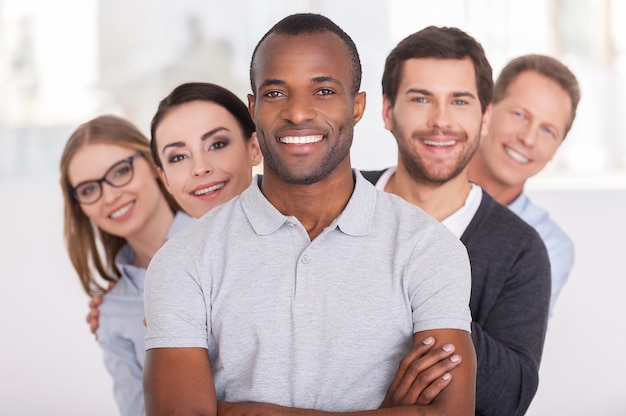 Equipo de negocios confiado. Alegre joven africano manteniendo los brazos cruzados y sonriendo mientras un grupo de personas de pie detrás de él en una fila y mirando a la cámara