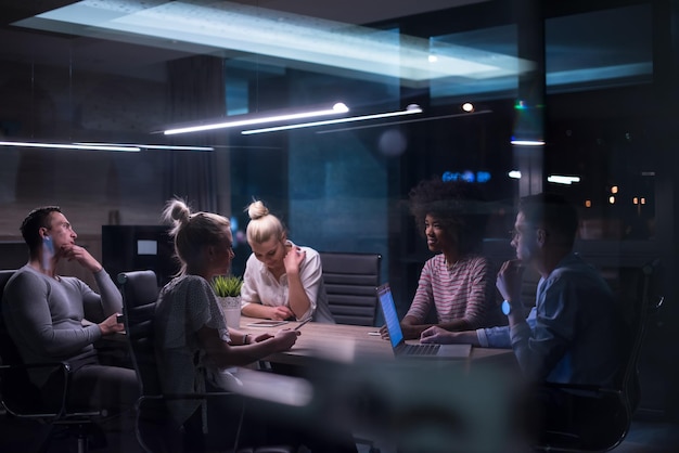 Equipo de negocios de inicio multiétnico en reunión en la lluvia de ideas interior de la oficina nocturna moderna, trabajando en la computadora portátil