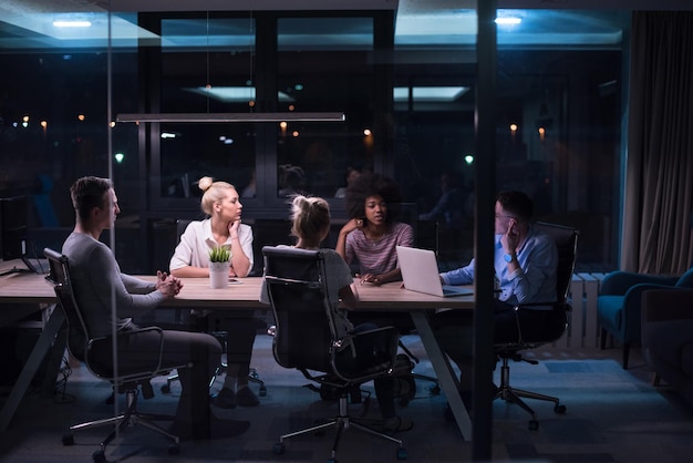 Equipo de negocios de inicio multiétnico en reunión en la lluvia de ideas interior de la oficina nocturna moderna, trabajando en la computadora portátil