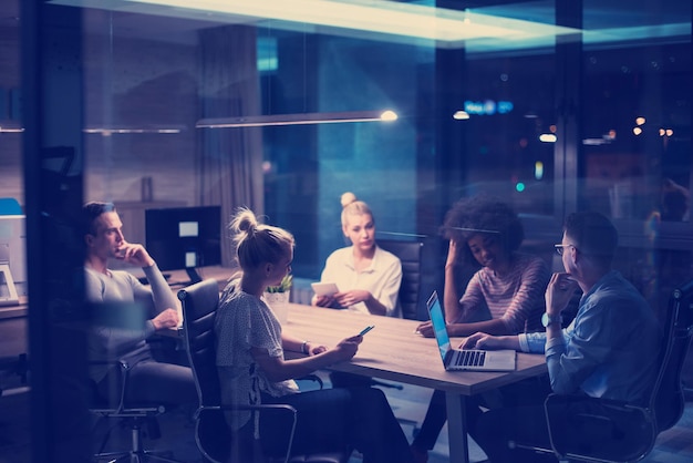 Equipo de negocios de inicio multiétnico en reunión en la lluvia de ideas interior de la oficina nocturna moderna, trabajando en la computadora portátil