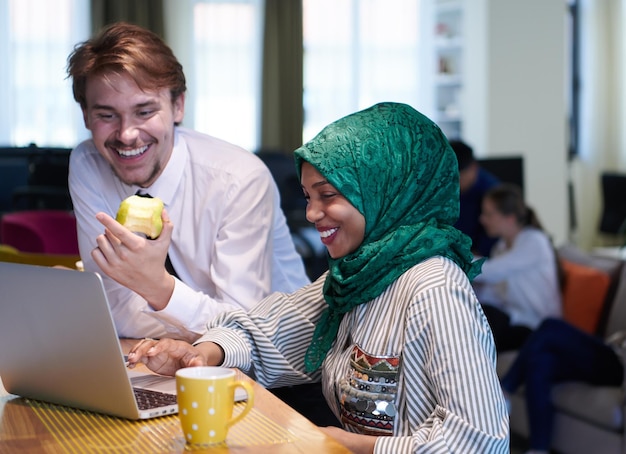 equipo de negocios multicultural internacional. hombre comiendo manzana mujer musulmana africana usando hiyab verde bebiendo té mientras trabajan juntos en una computadora portátil en el área de relajación en el moderno plan abierto startu