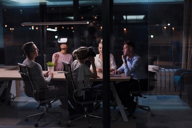 Equipo de negocios multiétnico que usa auriculares de realidad virtual en una reunión de oficina nocturna Reunión de desarrolladores con un simulador de realidad virtual alrededor de la mesa en una oficina creativa.