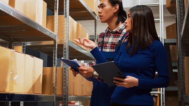 Equipo de personas asiáticas que verifican los suministros de existencias en tabletas y papeles, trabajando en el inventario de la sala de almacenamiento. Hombre y mujer jóvenes que usan dispositivos y archivos para el desarrollo de pequeñas empresas.