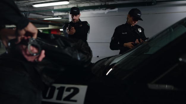 Foto equipo de policía arrestando a un criminal en un estacionamiento subterráneo policía masculino esposando a un sospechoso en un coche de patrulla oficial de policía apuntando a un ladrón con una pistola policía que usa una tableta digital para trabajar