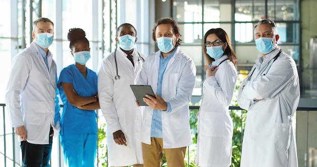 Equipo de razas mixtas de médicos profesionales masculinos y femeninos en el hospital. Grupo internacional de médicos en máscaras médicas con dispositivo de tableta Médicos multiétnicos en batas y uniformes en la clínica.