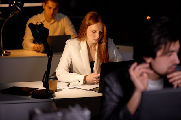 Equipo de trabajadores de oficina de gerentes que trabajan en el proyecto muy tarde, usando computadora. Personas trabajadoras en ropa formal que participan en el trabajo, el pensamiento, la lluvia de ideas, se centran en la mujer pelirroja con el teléfono inteligente