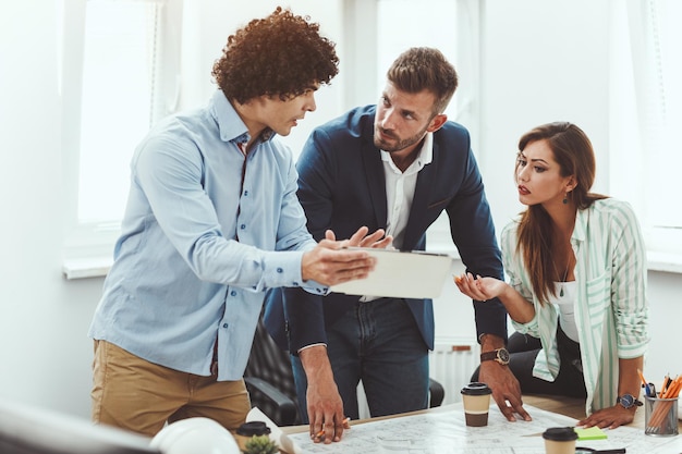 Equipo de tres jóvenes arquitectos que usan tabletas digitales y se comunican mientras analizan los planos de un nuevo proyecto en la oficina.