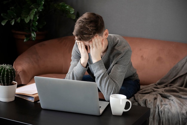 Er fühlt sich müde Frustrierter junger Mann, der erschöpft aussieht und sein Gesicht mit den Händen bedeckt, während er an seinem Laptop sitzt, seinem Arbeitsplatz zu Hause